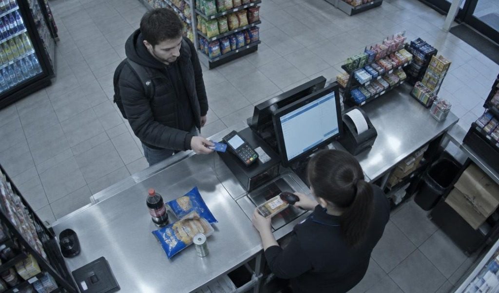 Overhead camera view of a checkout: a customer taps a card on the terminal while the cashier scans an item